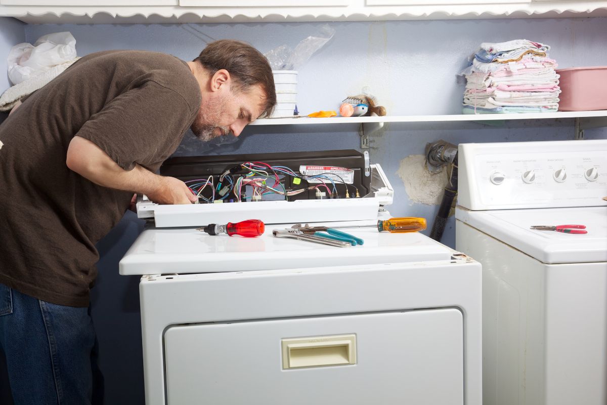  A man is repairing a dryer, focused on fixing the internal components with tools in hand.